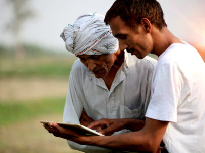 Young agronomist Standing during sunset near green field with senior Farmer holding tablet computer & showing something in the tablet computer to the farmer.