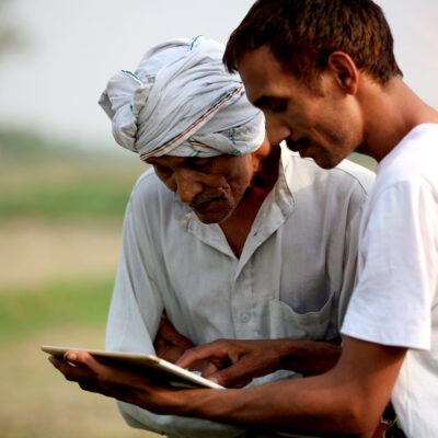 Young agronomist Standing during sunset near green field with senior Farmer holding tablet computer & showing something in the tablet computer to the farmer.
